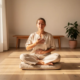 Persoon zit gekruist op linnen kussen in minimalistisch interieur, ogen gesloten, handen op borst en buik tijdens ademhalingsoefening.